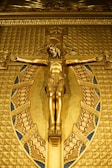 A golden depiction of Jesus on a crucifix, surrounded by an ornate oval frame with intricate patterns and Latin inscription. The artwork is rich in detail with a highly polished surface.