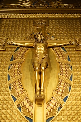 A golden depiction of Jesus on a crucifix, surrounded by an ornate oval frame with intricate patterns and Latin inscription. The artwork is rich in detail with a highly polished surface.