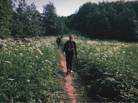 A woman walks along a narrow dirt path surrounded by lush green grass and white wildflowers on both sides. She carries a backpack and is dressed in dark clothing. The trail leads through a dense area of trees in the background, suggesting a rural or forest setting.