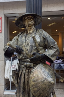 A person dressed in a detailed statue-like costume resembling a samurai, complete with traditional attire including a wide-brimmed hat and sword. The clothing and accessories appear to be made of materials that give them a weathered, bronze-like appearance. This figure is standing outside, possibly near a store, as evidenced by the clothing items visible in the background display.