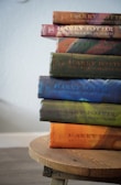 A stack of hardbound Harry Potter books arranged on a wooden stool. The titles visible include 'Harry Potter and the Sorcerer's Stone', 'Harry Potter and the Chamber of Secrets', 'Harry Potter and the Prisoner of Azkaban', 'Harry Potter and the Goblet of Fire', 'Harry Potter and the Order of the Phoenix', 'Harry Potter and the Half-Blood Prince', and 'Harry Potter and the Deathly Hallows'. The books are in different colors, and are arranged in order with the first book on top.