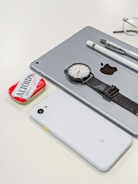 A collection of personal items including a tablet with an Apple logo, a smartwatch with a black strap, a white smartphone with a Google logo, a white stylus, a pen, and a red tin of Altoids mints. Glasses are partially visible in the background.
