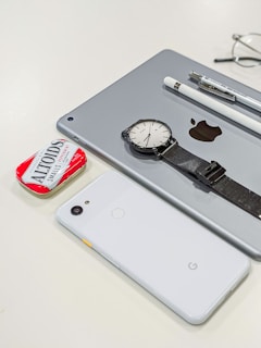 A collection of personal items including a tablet with an Apple logo, a smartwatch with a black strap, a white smartphone with a Google logo, a white stylus, a pen, and a red tin of Altoids mints. Glasses are partially visible in the background.