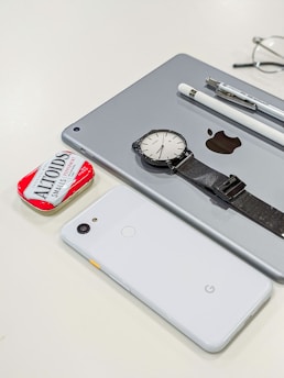 A collection of personal items including a tablet with an Apple logo, a smartwatch with a black strap, a white smartphone with a Google logo, a white stylus, a pen, and a red tin of Altoids mints. Glasses are partially visible in the background.