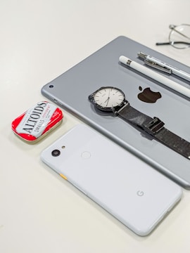 A collection of personal items including a tablet with an Apple logo, a smartwatch with a black strap, a white smartphone with a Google logo, a white stylus, a pen, and a red tin of Altoids mints. Glasses are partially visible in the background.