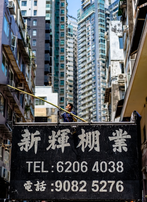A person standing on a high platform with a long pole, surrounded by tall residential buildings. An advertisement board with phone numbers and Asian script appears in the foreground, partially obscuring the person's lower body.