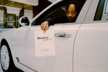 A person is seated in the driver’s seat of a white luxury car, holding a paper sign that reads 'Bentley Records' out of the window. The vehicle is inside a well-lit showroom with a modern architectural design.