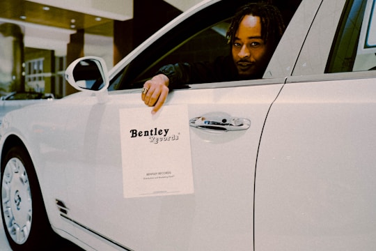 A person is seated in the driver’s seat of a white luxury car, holding a paper sign that reads 'Bentley Records' out of the window. The vehicle is inside a well-lit showroom with a modern architectural design.
