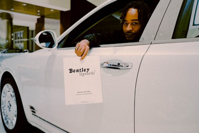 A person is seated in the driver’s seat of a white luxury car, holding a paper sign that reads 'Bentley Records' out of the window. The vehicle is inside a well-lit showroom with a modern architectural design.