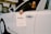 A person is seated in the driver’s seat of a white luxury car, holding a paper sign that reads 'Bentley Records' out of the window. The vehicle is inside a well-lit showroom with a modern architectural design.