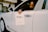A person is seated in the driver’s seat of a white luxury car, holding a paper sign that reads 'Bentley Records' out of the window. The vehicle is inside a well-lit showroom with a modern architectural design.