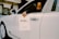 A person is seated in the driver’s seat of a white luxury car, holding a paper sign that reads 'Bentley Records' out of the window. The vehicle is inside a well-lit showroom with a modern architectural design.