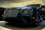 A luxury car is displayed in a showroom with a sleek design and glossy finish. The front grille is prominent, with intricate mesh detailing and chrome accents. The headlights are modern and stylish, and the car's body reflects the showroom lights, adding to its elegance.