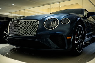 A luxury car is displayed in a showroom with a sleek design and glossy finish. The front grille is prominent, with intricate mesh detailing and chrome accents. The headlights are modern and stylish, and the car's body reflects the showroom lights, adding to its elegance.