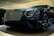 A luxury car is displayed in a showroom with a sleek design and glossy finish. The front grille is prominent, with intricate mesh detailing and chrome accents. The headlights are modern and stylish, and the car's body reflects the showroom lights, adding to its elegance.