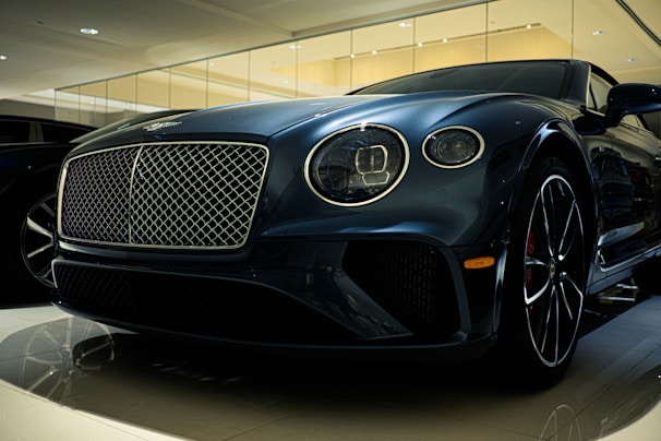 A luxury car is displayed in a showroom with a sleek design and glossy finish. The front grille is prominent, with intricate mesh detailing and chrome accents. The headlights are modern and stylish, and the car's body reflects the showroom lights, adding to its elegance.