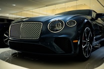 A luxury car is displayed in a showroom with a sleek design and glossy finish. The front grille is prominent, with intricate mesh detailing and chrome accents. The headlights are modern and stylish, and the car's body reflects the showroom lights, adding to its elegance.