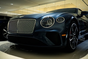 A luxury car is displayed in a showroom with a sleek design and glossy finish. The front grille is prominent, with intricate mesh detailing and chrome accents. The headlights are modern and stylish, and the car's body reflects the showroom lights, adding to its elegance.