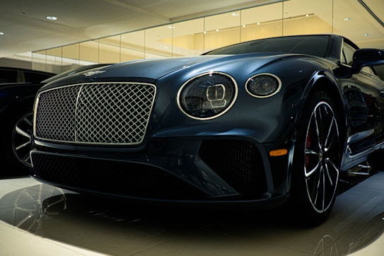 A luxury car is displayed in a showroom with a sleek design and glossy finish. The front grille is prominent, with intricate mesh detailing and chrome accents. The headlights are modern and stylish, and the car's body reflects the showroom lights, adding to its elegance.