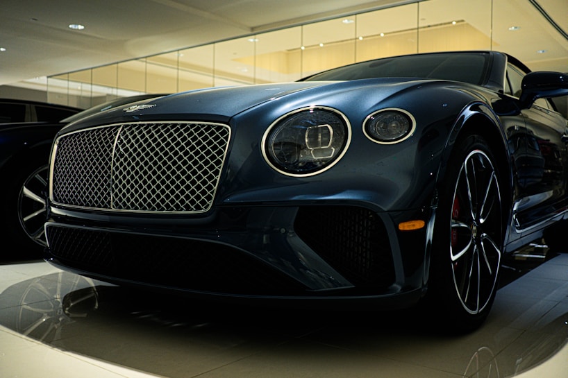 A luxury car is displayed in a showroom with a sleek design and glossy finish. The front grille is prominent, with intricate mesh detailing and chrome accents. The headlights are modern and stylish, and the car's body reflects the showroom lights, adding to its elegance.