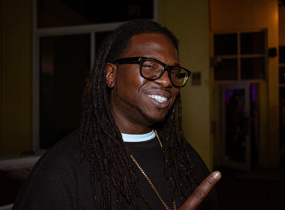 Man with long dreadlocks stands in front of a yellow wall wearing glasses and smile