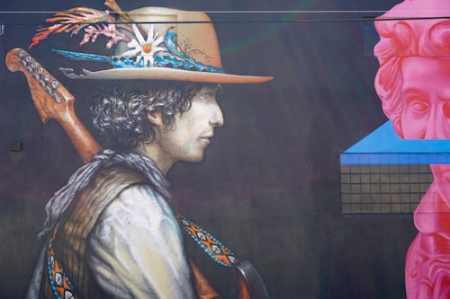 A mural depicting a person in profile, wearing a large, decorated hat with feathers and floral designs. The individual has curly hair and is carrying a stringed instrument over their shoulder. The mural includes vibrant colors and detailed patterns on the person's clothing. To the right, there is a large, pink classical statue head set against a modern, geometric shape with a grid pattern.