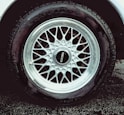 A detailed view of a car tire with an intricate metallic alloy wheel design, featuring multiple interlocking spokes. The brand name is visible on the wheel cap against the dark rubber of the tire. The wheel is partially wet, likely from rain or washing, with droplets visible.