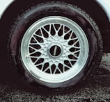 A detailed view of a car tire with an intricate metallic alloy wheel design, featuring multiple interlocking spokes. The brand name is visible on the wheel cap against the dark rubber of the tire. The wheel is partially wet, likely from rain or washing, with droplets visible.