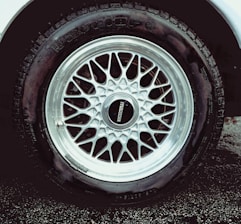 A detailed view of a car tire with an intricate metallic alloy wheel design, featuring multiple interlocking spokes. The brand name is visible on the wheel cap against the dark rubber of the tire. The wheel is partially wet, likely from rain or washing, with droplets visible.