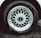 A detailed view of a car tire with an intricate metallic alloy wheel design, featuring multiple interlocking spokes. The brand name is visible on the wheel cap against the dark rubber of the tire. The wheel is partially wet, likely from rain or washing, with droplets visible.