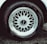 A detailed view of a car tire with an intricate metallic alloy wheel design, featuring multiple interlocking spokes. The brand name is visible on the wheel cap against the dark rubber of the tire. The wheel is partially wet, likely from rain or washing, with droplets visible.