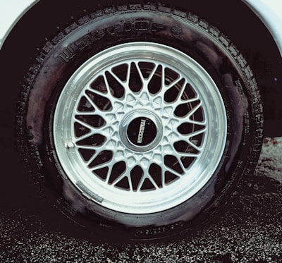 A detailed view of a car tire with an intricate metallic alloy wheel design, featuring multiple interlocking spokes. The brand name is visible on the wheel cap against the dark rubber of the tire. The wheel is partially wet, likely from rain or washing, with droplets visible.