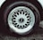A detailed view of a car tire with an intricate metallic alloy wheel design, featuring multiple interlocking spokes. The brand name is visible on the wheel cap against the dark rubber of the tire. The wheel is partially wet, likely from rain or washing, with droplets visible.