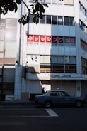 A city street scene with a light blue taxi parked on the roadside. Behind it, a building facade features large windows and a sign with Japanese text in red and white, along with another sign reading 'Natural Greens.' The foreground includes overhanging branches with green leaves casting shadows on the building.