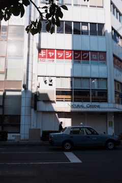 A city street scene with a light blue taxi parked on the roadside. Behind it, a building facade features large windows and a sign with Japanese text in red and white, along with another sign reading 'Natural Greens.' The foreground includes overhanging branches with green leaves casting shadows on the building.