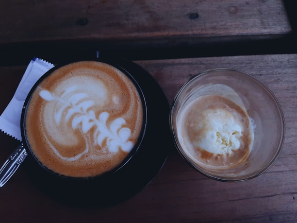 A frothy cappuccino with intricate latte art is placed next to an affogato dessert, with a scoop of vanilla ice cream partly submerged in espresso. Both are set on a wooden table, accompanied by a spoon and a small napkin.