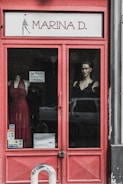 A storefront display with a red-framed glass door. Visible are mannequins dressed in elegant gowns, one in a red dress and the other in a black one. The name 'Marina D.' is prominently displayed above the door. Sticker labels for payment options are affixed to the door, and a car is partially visible reflected in the glass.