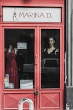 A storefront display with a red-framed glass door. Visible are mannequins dressed in elegant gowns, one in a red dress and the other in a black one. The name 'Marina D.' is prominently displayed above the door. Sticker labels for payment options are affixed to the door, and a car is partially visible reflected in the glass.