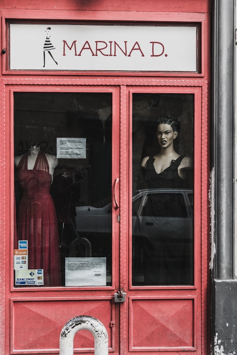 A storefront display with a red-framed glass door. Visible are mannequins dressed in elegant gowns, one in a red dress and the other in a black one. The name 'Marina D.' is prominently displayed above the door. Sticker labels for payment options are affixed to the door, and a car is partially visible reflected in the glass.