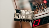 A close-up view of an electronic component labeled with WiFi capability, specifically the ESP8266MOD. The component has intricate circuit patterns and is situated near a digital display showing the number 17. The background includes blurry wiring elements and a slight red light glow, suggesting an electronic device setup.