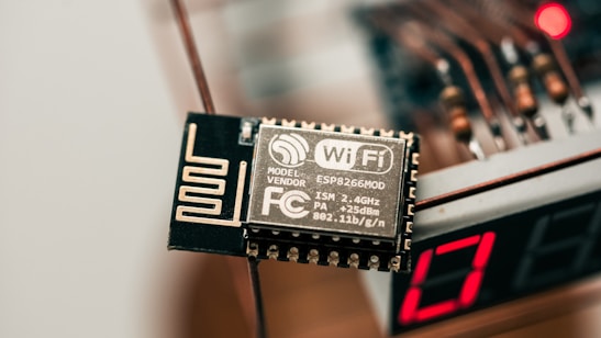 A close-up view of an electronic component labeled with WiFi capability, specifically the ESP8266MOD. The component has intricate circuit patterns and is situated near a digital display showing the number 17. The background includes blurry wiring elements and a slight red light glow, suggesting an electronic device setup.