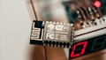 A close-up view of an electronic component labeled with WiFi capability, specifically the ESP8266MOD. The component has intricate circuit patterns and is situated near a digital display showing the number 17. The background includes blurry wiring elements and a slight red light glow, suggesting an electronic device setup.
