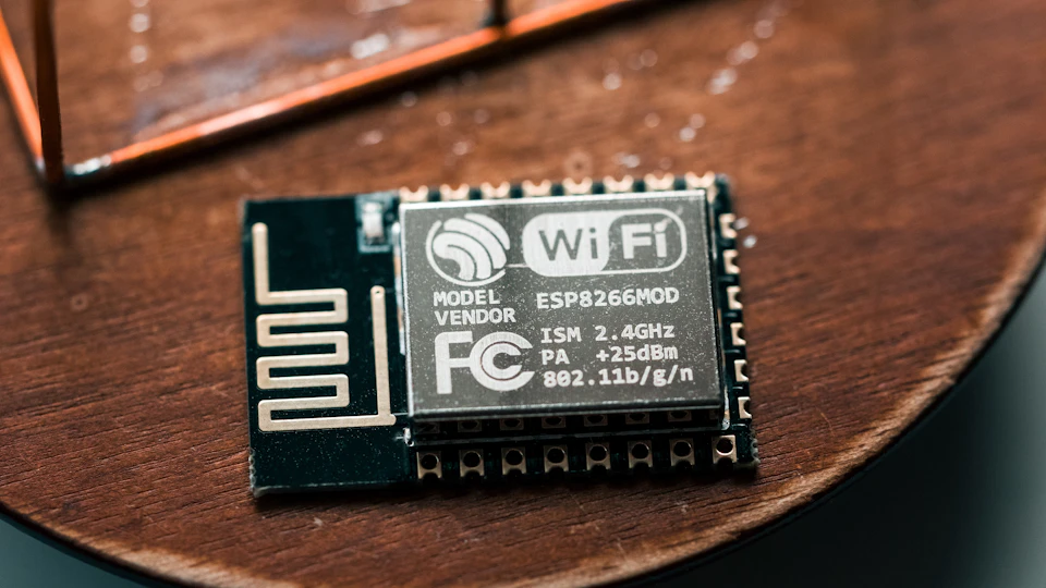 The image shows a small black and silver electronic device, likely a component or circuit board, resting on a wooden surface. The device is placed on its side with the top facing upwards, giving it a three-dimensional appearance. The device appears to be a model of an ESP8266, which is a type of microcontroller used in various applications such as robotics and automation. The ESP8266 has a white label on its back that displays the text "Model Vender" and "ISH 2.4 Ghz".