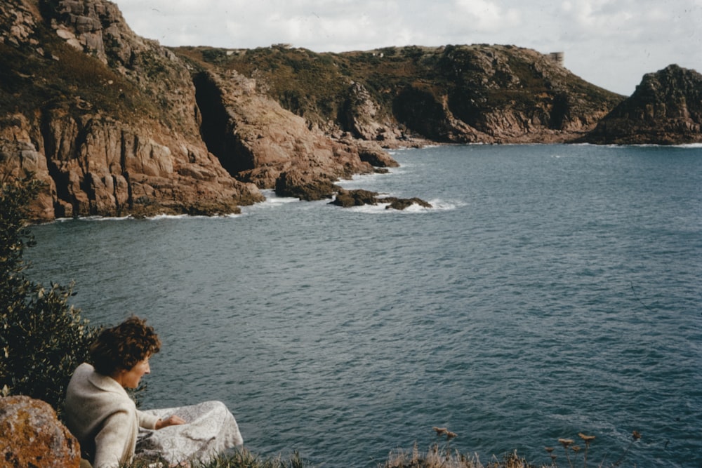 woman in white long sleeve shirt sitting on rock formation near body of water during daytime