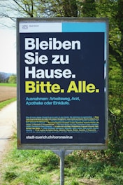 A public sign in multiple languages urging people to stay at home, with exceptions for work, medical appointments, and essential shopping. The text is prominently displayed in bold white and yellow font on a dark background, and the sign is located outdoors next to a path and greenery.