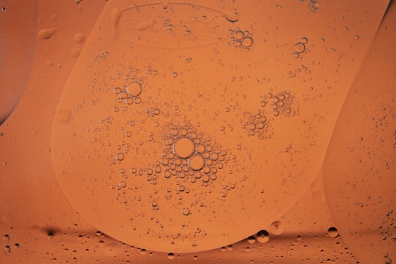 A close-up view of oil droplets suspended in a liquid, creating a pattern of small and large circles on an orange background. The droplets are varied in size and appear to cluster together in certain areas, giving a textured appearance.