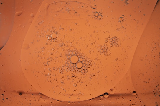 A close-up view of oil droplets suspended in a liquid, creating a pattern of small and large circles on an orange background. The droplets are varied in size and appear to cluster together in certain areas, giving a textured appearance.