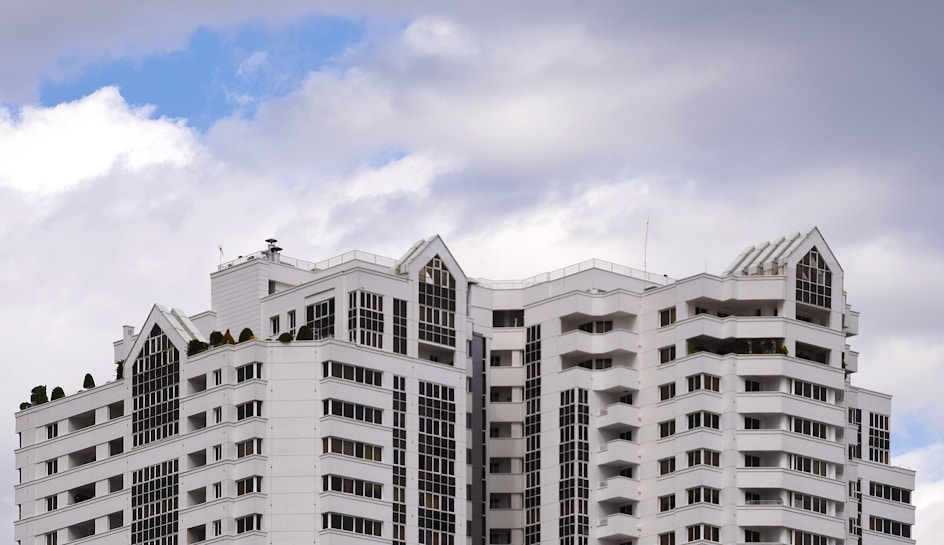 A modern high-rise building with white and light grey exterior, featuring multiple balconies and large windows. The architectural design includes sharp angles and geometric patterns. The background is filled with a partly cloudy sky, with patches of blue visible.