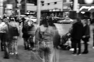 A bustling city street with blurred motion capturing pedestrians, suggesting movement and a busy atmosphere. Buildings and parked cars are visible in the background, adding to the urban setting.