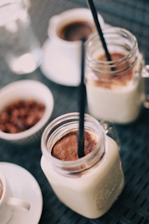 Several beverages, including two creamy drinks in mason jars with black straws and some coffee cups, are arranged on a mat. There is also a small bowl filled with what looks like chocolate or nuts.